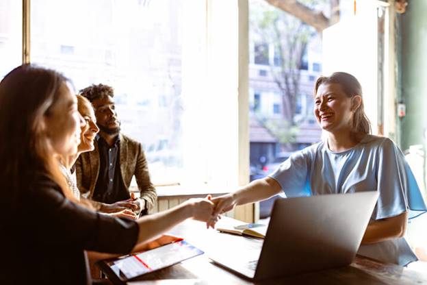 A diverse group of people are meeting at a table. A woman shakes hands with another while a laptop is visible.